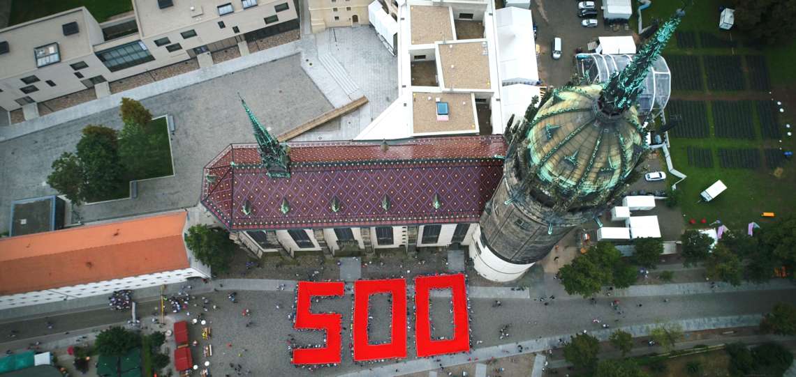Luftaufnahme der Schlosskirche Wittenberg, vor der viele Menschen zusammen eine rote 500 aus Stoff halten