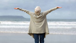 Ältere Frau steht mit ausgebreiteten Armen am Strand