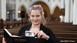 Eine Frau Mitte 20 sitzt in einer Kirchenbank und lächelt in die Kamera, in einer Hand eine Leuchtlupe, in der anderen eine Bibel.