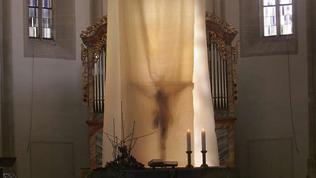 Morgenlicht scheint durch ein Tuch, welches das Altarkreuz in der evangelischen Michaelskirche in Ammerbuch-Entringen bei Tübingen bis zum Karfreitag verhüllt.