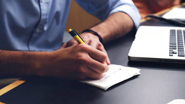 Ein Mann sitzt vor einem Laptop und macht sich handschriftliche Notizen.