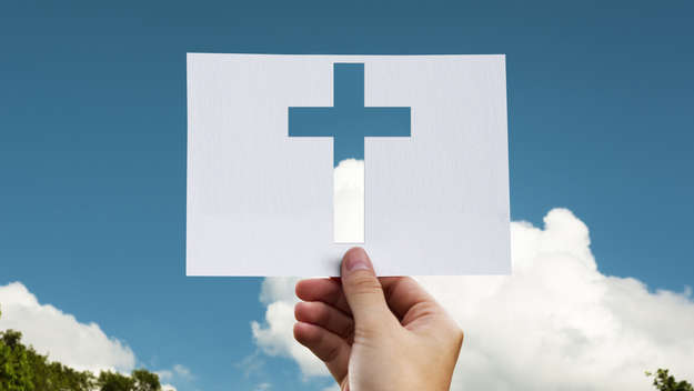 Hand mit einem Blatt Papier, aus dem ein Kreuz ausgeschnitten wurde.