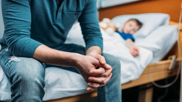 Ein Mann sitzt mit gefalteten Händen auf dem Krankenhausbett eines Kindes.