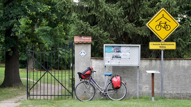 Bepacktes Fahrrad lehnt an der Mauer eines Kirchhofs neben dem Hinweisschild mit der Aufschrift '1. Deutsche Radfahrerkirche Wessnig' am Elbe-Radweg (Sachsen)