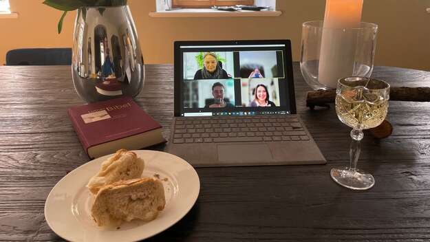 Man sieht einen Tisch mit einem aufgeklappten Notebook und Teilnehmenden einer Zoom Konferenz, daneben einen Teller mit einem Stück Brot, ein gefülltes Weinglas, eine angezündete Kerze und eine Bibel