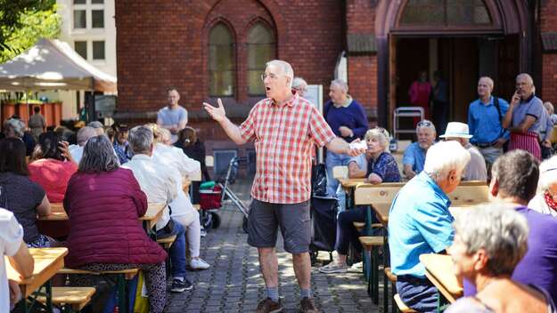 Pastor Uwe Sassnowski (Saßnowski) beim 'Mittagstisch fuer alle' an der Evangelisch-methodistischen Christuskirche am Merianplatz in Frankfurt am Main Um ihn herum sitzen Menschen an Bierzeltgarnituren und essen ihr Mittagessen.