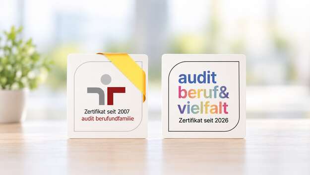 Zwei Zertifikatslogs stehen nebeneinander auf einem hellen Holztisch vor weich verschwommenem Hintergrund: audit berufundfamilie', zertifiziert seit 2007, und audit beruf&vielfalt, zertifiziert seit 2026