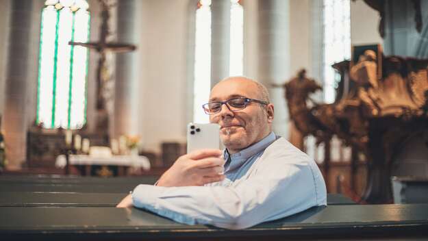 Zu sehen ist Pfarrer Lutz Neumeier. Er sitzt in seiner Alltagskleidung in der Kirchenbank. In der rechten Hand hält er sein weißes Smartphone