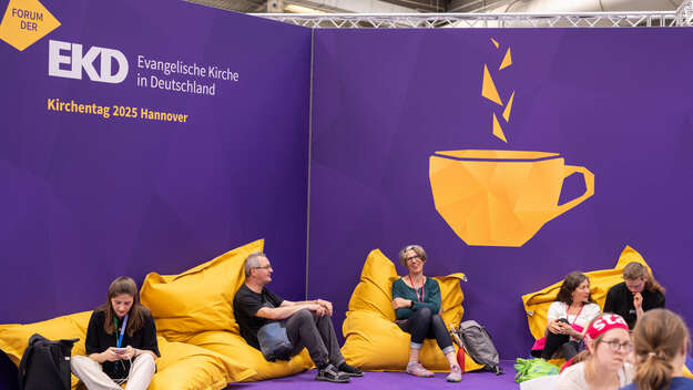 Besucher ruhen sich auf dem Forum der EKD beim Kirchentag auf gelben Sitzsäcken aus vor einer lila Rückwand