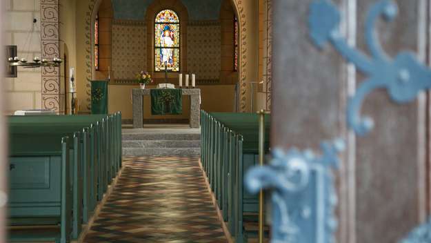 Im Vordergrund rechts die Eingangstür zur Kirche. Bunte Bodenfliesen, Verzierungen am Bogen. Helle Fenster, im Altarraum hellgrauer Steinboden.