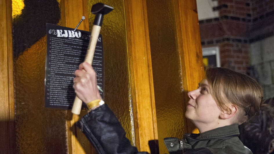 Junge Frau schlägt Thesenpapier mit einem Hammer an Kirchwand