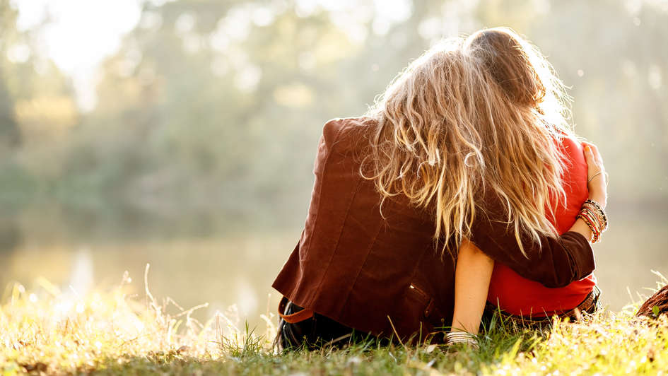 Zwei junge Frauen sitzen Arm in Arm auf einer Wiese.