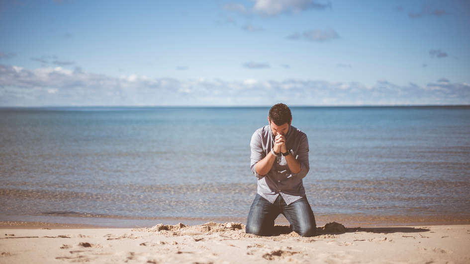 Junger Mann kniet betend am Strand