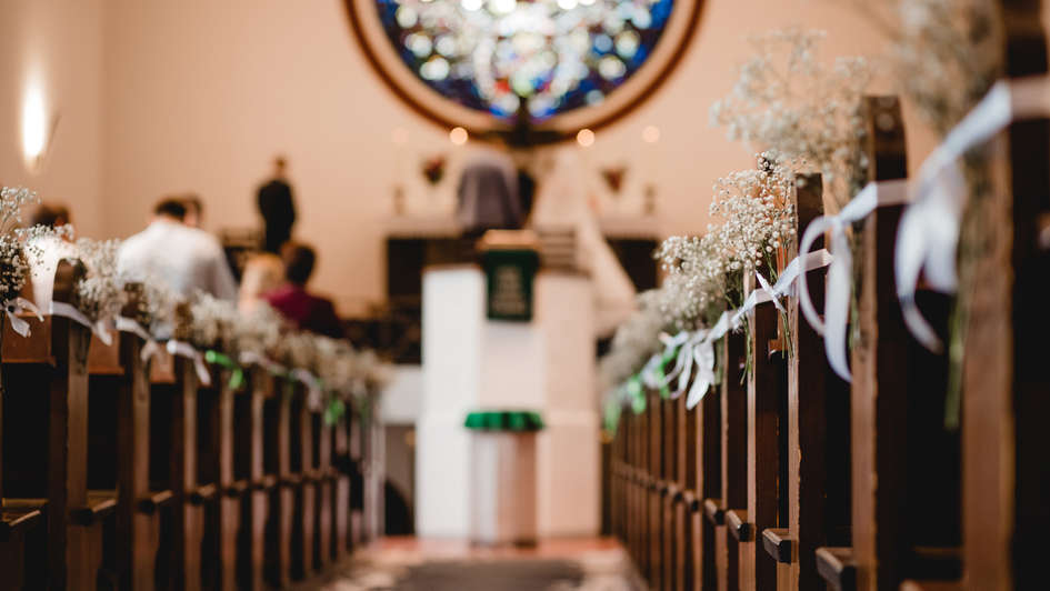 Blick durch den Mittelgang einer geschmückten Kirche, vorm Altar steht ein Brautpaar