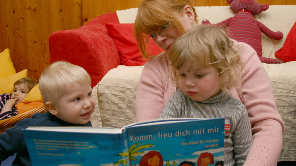 Eine Erzieherin hat zwei beiden Zweijährige auf dem Schoß und liest ihnen aus einer Kinderbeibel vor Eine Erzieherin hat zwei beiden Zweijährige auf dem Schoß und liest ihnen aus einer Kinderbeibel vor