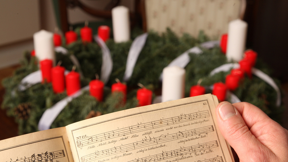Liederbuch 'Unsere Lieder' der 'Agentur des Rauhen Hauses' in Hamburg von 1844, mit dem Weihnachtslied 'Stille Nacht, heilige Nacht', im Hintergrund ein Wichern-Adventskranz