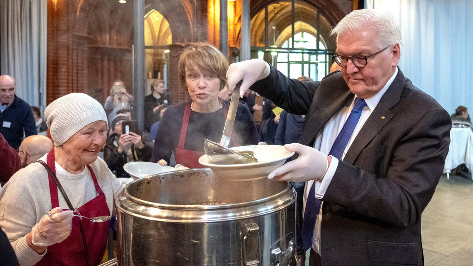 Bundespräsident Frank-Walter Steinmeier füllt mit einer Suppenkelle Suppe aus einem großen Kessel in einen Teller, seine Frau Elke Büdenbender steht in Schürze dabei.