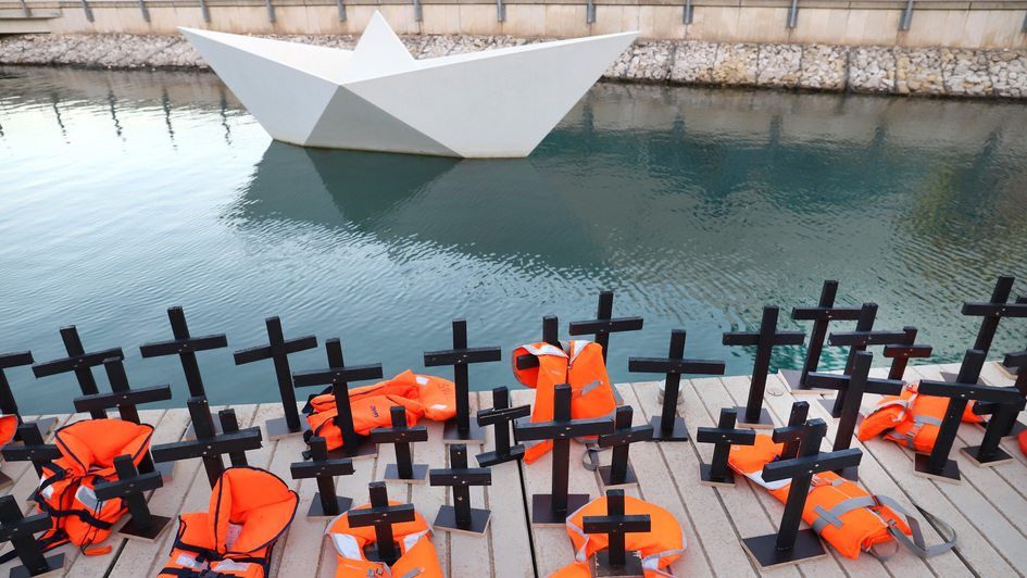 Kreuze und Schwimmwesten auf einem Steg, ein überdimensionales Papierboot im Wasser: Szene bei einer Gedenkfeier für ertrunkene Flüchtlinge im Hafen von Valletta (Malta).