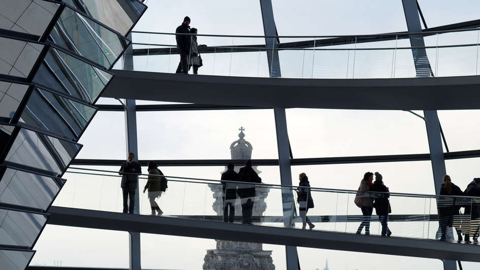 Blick aus der gläsernen Kuppel des Reichstagsgebäudes, in der Besucher herumlaufen, auf ein Türmchen des Gebäudes, auf dem ein Kreuz steht.