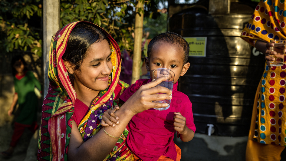 Eine Mutter in Bangladesch hält ihr Kleinkind auf dem Arm und gibt ihm ein Glas Wasser zu trinken