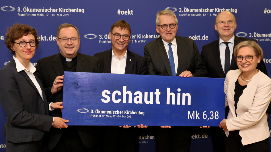 Verantowrtliche des Ökumenischen Kirchentags 2021 in Frankfurt am Main halten eine Tafel mit dem Leitwort der Veranstaltung: 'Schaut hin' (Mk 6,38)