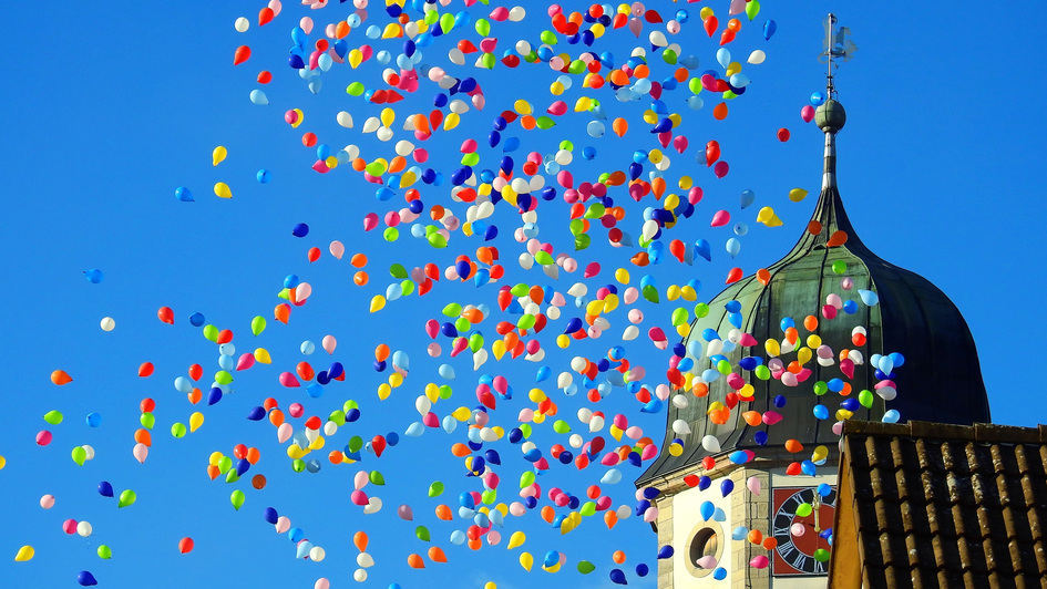 Vor einem Kirchturm steigen hunderte bunter Luftballons in den blauen Himmel