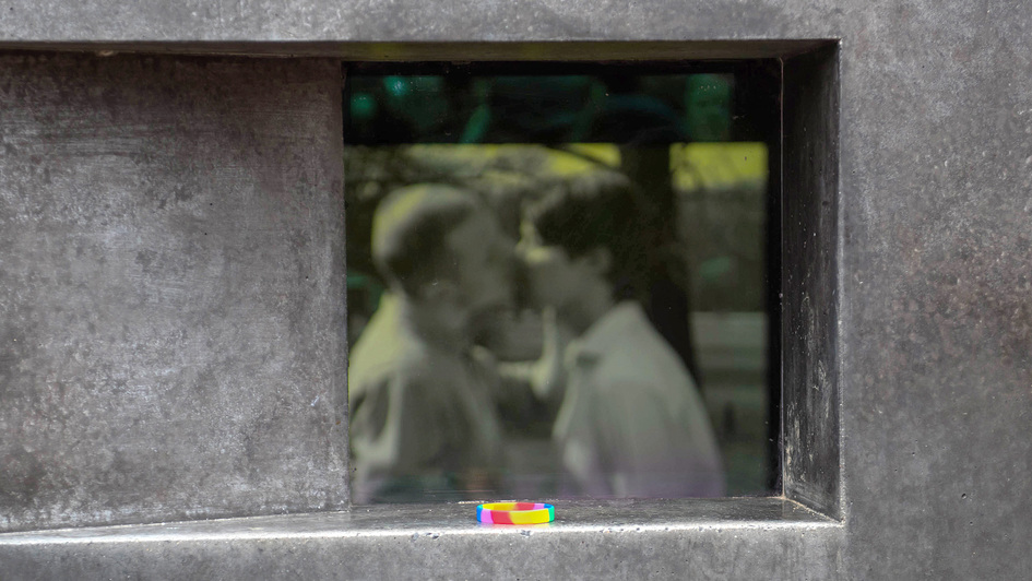 Regenbogen-Armband am Denkmal für die im Nationalsozialismus verfolgten Homosexuellen in Berlin-Tiergarten. Im Hintergrund läuft ein Film mit einer Szene, in der sich zwei Männer küssen.