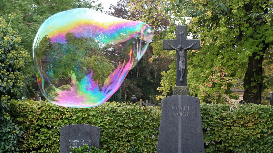 Eine große schillernde Seifenblase schwebt zwischen Grabsteinen auf einem Friedhof. Eine große schillernde Seifenblase schwebt zwischen Grabsteinen auf einem Friedhof.