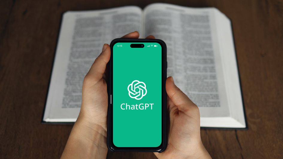 Eine Person hält ein Smartphone mit dem ChatGPT-Logo auf grünem Bildschirm in den Händen; im Hintergrund liegt ein geöffnete Bibel auf einem Holztisch.