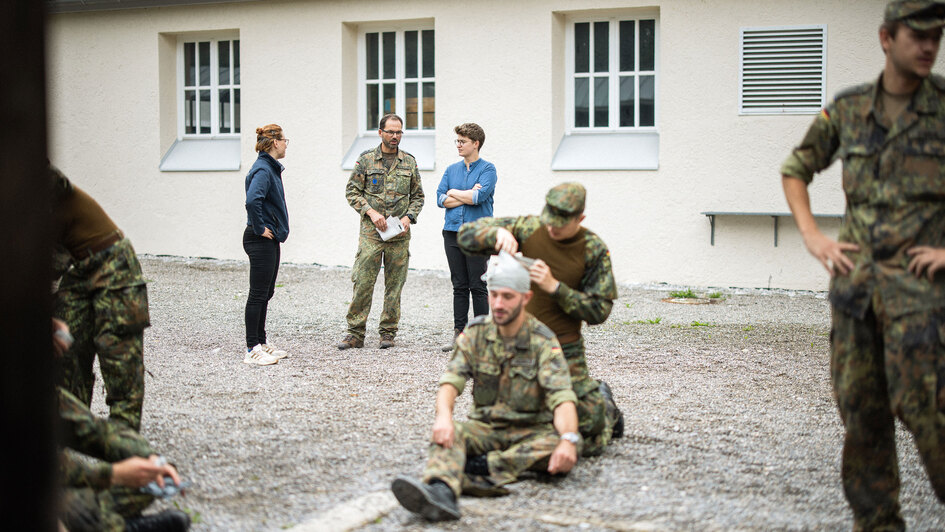 Anna-Nicole Heinrich im Gespräch mit einem Bundeswehrsoldaten während ihrer Präsestour auf einem Bundeswehrstützpunkt. Im Vordergrund üben Soldaten die Anlage eines Kopfverbandes, weitere Soldaten stehen und beobachten die Situation