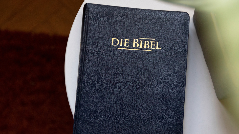 Eine schwarze Bibel mit goldener Aufschrift „DIE BIBEL“ liegt auf einem weißen Tisch. Der Einband wirkt leicht strukturiert, und im Hintergrund ist ein unscharfer roter Teppich zu erkennen.
