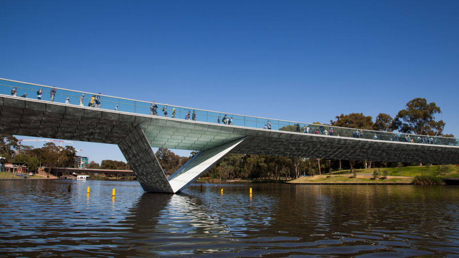 Moderne Fußgängerbrücke mit gläsernem Geländer über einen Fluss, zahlreiche Menschen überqueren die Brücke bei blauem Himmel, grüne Parkanlage im Hintergrund.