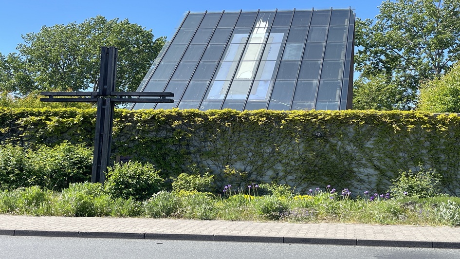Zu sehen ist die Autobahnkirche Medenbach. Das große Glasdach ragt hinter einer Steinmauer hervor. Vor der Mauer steht ein großes schwarzes Stahlkreuz