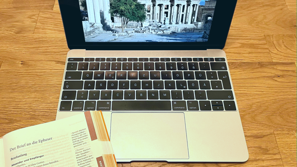Aufgeklappter Laptop mit einer Bibel, die neben der Tastatur liegt. Auf dem Bildschirm ist ein Bild von Ruinen mir griechischen Säulen zu sehen. Die Bibel sit beim Epheserbrief aufgeschlagen.