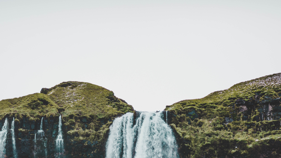 Oberer Teil eines Wasserfalls von einem begrüntem Hügel mit grauem Himmel.