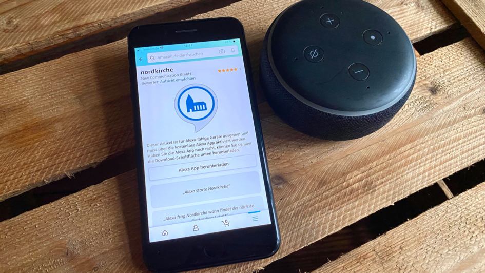 Ein Smartphone und ein Lautsprecher-Sprachassistent liegen auf einer Tischplatte. Auf dem Smartphone ist Amazon geöffnet und man sieht einen Skill mit dem Namen 'nordkirche' und ein blaues Piktogramm von einer Kirche in einem Kreis.