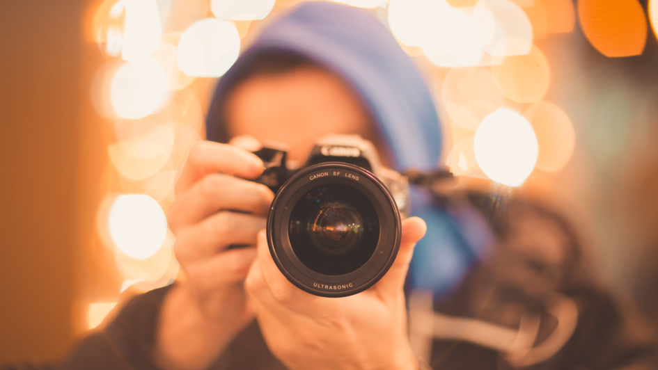 Mensch blauer Kapuze und mit Kamera vor dem Gesicht. Er sieht aus, als würde er ein Foto direkt in die Kamera, die ihn fotografiert schießen. Der Fokus ist auf die Kamera gerichtet. Der Mensch und der Hintergrund sind unscharf. Mensch blauer Kapuze und mit Kamera vor dem Gesicht. Er sieht aus, als würde er ein Foto direkt in die Kamera, die ihn fotografiert schießen. Der Fokus ist auf die Kamera gerichtet. Der Mensch und der Hintergrund sind unscharf.