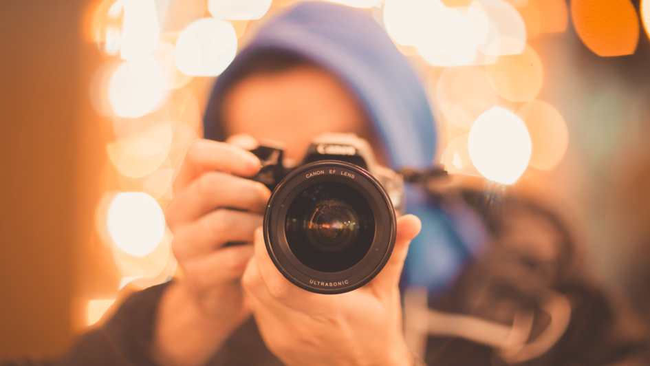 Mensch blauer Kapuze und mit Kamera vor dem Gesicht. Er sieht aus, als würde er ein Foto direkt in die Kamera, die ihn fotografiert schießen. Der Fokus ist auf die Kamera gerichtet. Der Mensch und der Hintergrund sind unscharf.