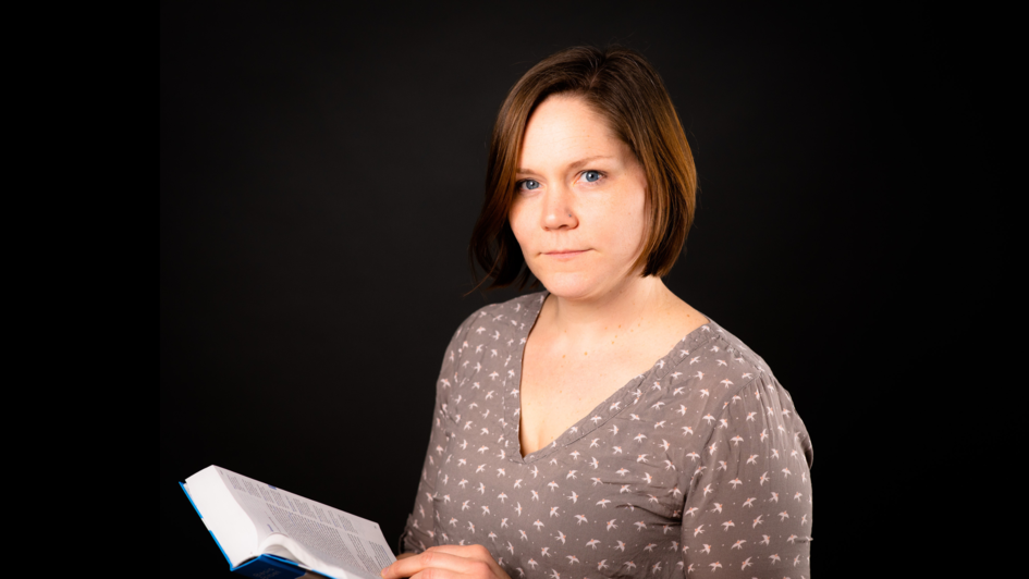 Weibliche erson mittleren Alters (Pfarrerin Meike Melchinger) mit einem Buch in der Hand vor schwarzem Hintergrund.