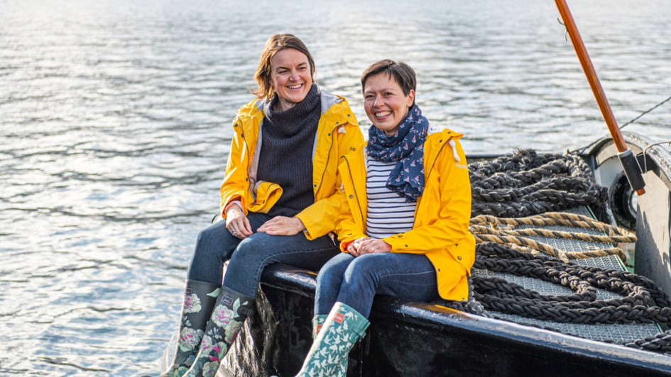 Zwei Frauen (Nadia Kamoun und Claudia Aue) sitzen auf einem Kai. Sie tragen eine gelbe Regenjacke und Gummistiefel. Hinter ihnen liegen Taue, im Hintergrund ist Wasser zu sehen.