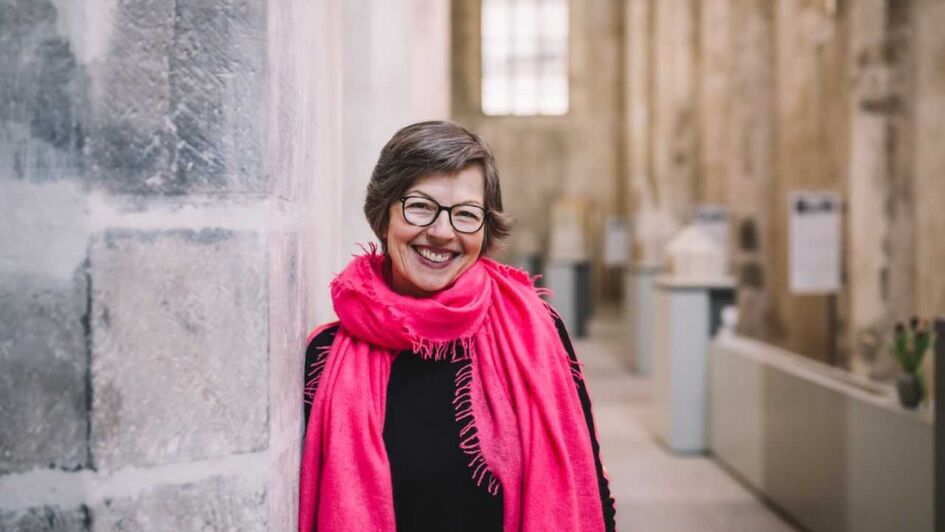 Catharina Hasenclever, traegt kurze braune Haare. Sie lacht in die Kamera. Um ihren Hals liegt ein pinker Schal. Sie lehnt an einer Kirchenmauer