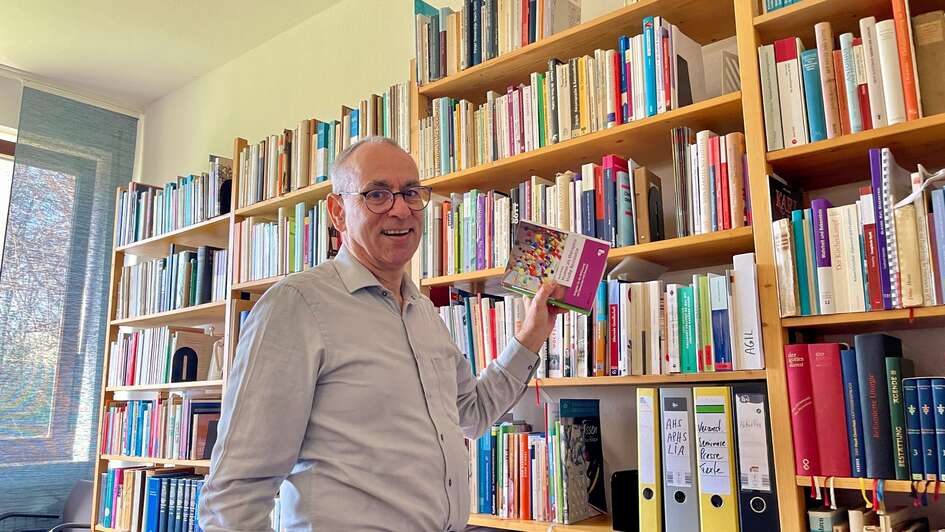 Zu sehen ist der Pfarrer und Seelsorger Christian Wiener. Er trägt Brille und ein karriertes Hemd. Im Halbprofil blickt er in die Kamera. Er steht an einem vollen Buchregal und hat ein Buch in der Hand