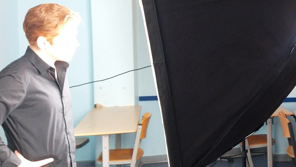 Person in schwarzem Hemd steht bei einem Lichttest in einem Klassenzimmer vor einem großen Softbox-Studiolicht. Im Hintergrund sind leere Tische und Stühle zu sehen.