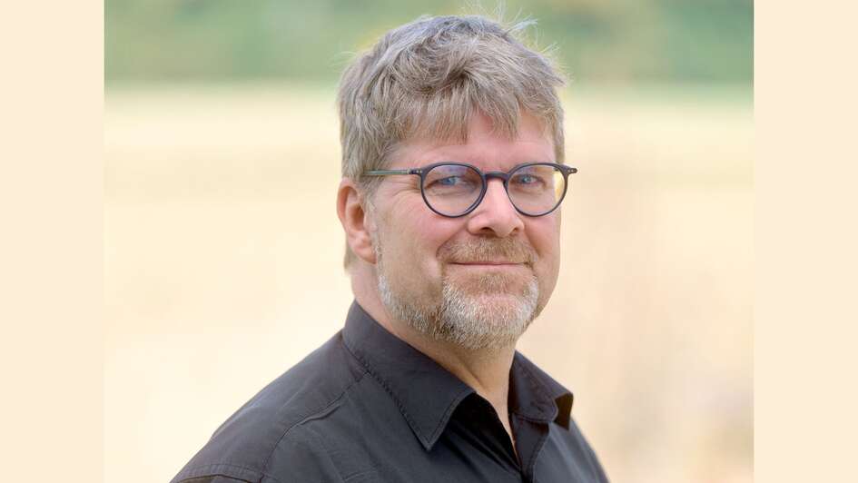 Eine seitliche portraitaufnahme von Dr. Fabian Vogt. Er trägt hellbraunes kurzes Haat, dass ihm auch ein Stück in die Stirn ragt, eine dunkle, runde Brille und einen gräulich-braunenDreitage Bart. Sein Hemd ist schwarz, der obere Knopf ist geöffnet.