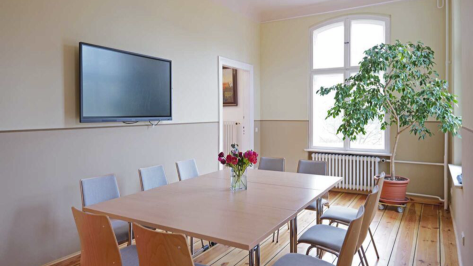 Heller Seminarraum mit großen Fenstern, einem rechteckigen Tisch, umliegenden Stühlen, einem Wandbildschirm, einem Strauß Blumen in einer Vase und einer großen Zimmerpflanze in der Ecke.