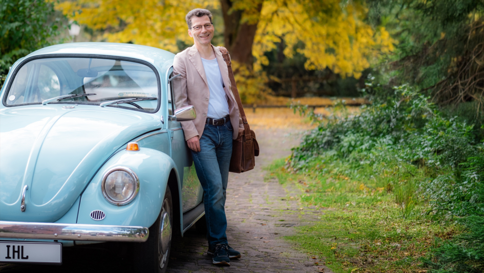 Prof. Dr. Christoph Schrodt steht lächelnd mit Ledertasche neben einem hellblauen VW-Käfer auf einem Waldweg. Das Auto symbolisiert das in der Predigtwerkstatt verwendete Modell des „Predigtautos“.