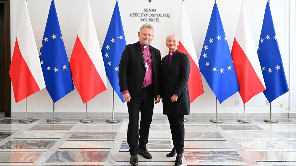 Im Gebäude des Senats der Republik Polen stehen Bischöfin Kirsten Fehrs (rechts) und Bischof Jerzy Samiec (links) nebeneinander. Beide tragen schwarze Kleidung mit violettem Hemd und Bischofskreuz. Hinter ihnen sind abwechselnd polnische und EU-Flaggen so