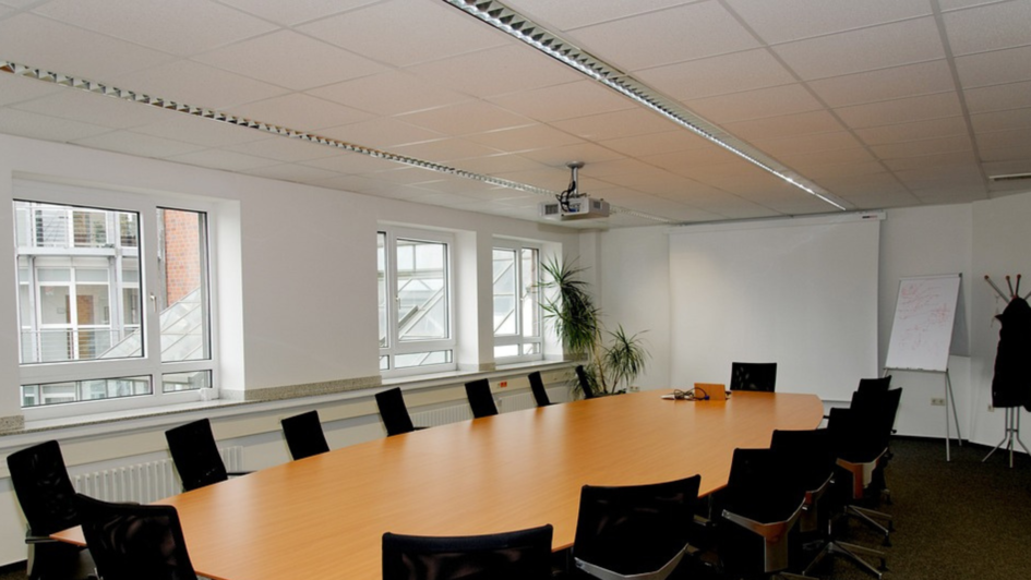Das Bild zeigt einen leeren Konferenzraum. In der Mitte befindet sich ein langer Tisch mit Bürostühlen drumherum.  An einer Wand befindet sich eine Leinwand; auf diese ist ein Beamer gerichtet, der an der Decke befestigt wurde.  Neben der Leinwand stehen