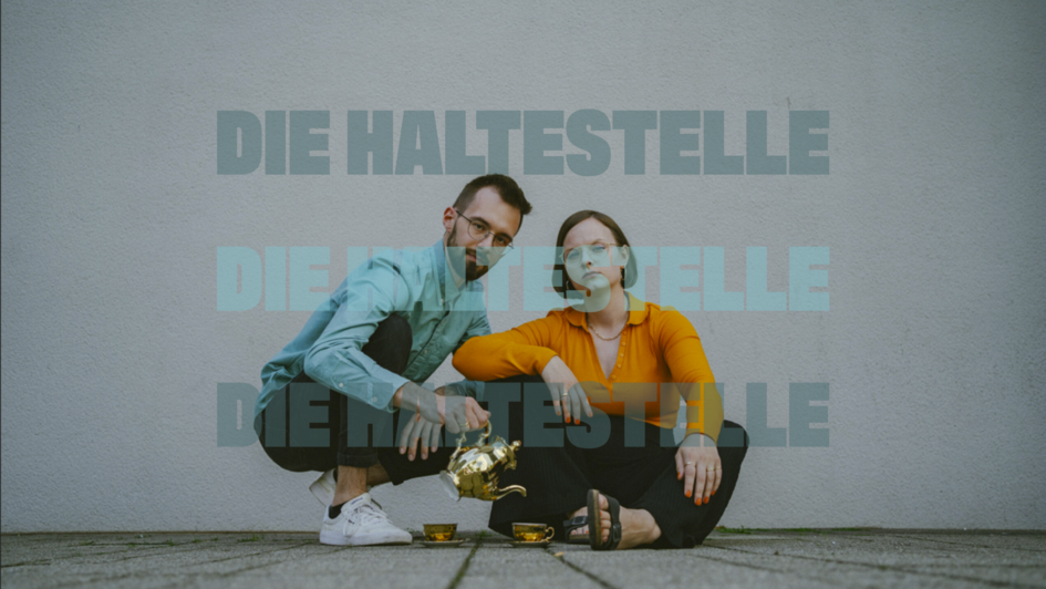 Zwei  Erwachsene sitzen nebeneinander auf dem Boden vor einer grauen Wand. Sie schauen ernst in die Kamera. Die Person links trägt ein türkisfarbenes Hemd, weiße Sneaker und schenkt mit einer goldenen Teekanne Tee in eine kleine Tasse. Die Person rechts t