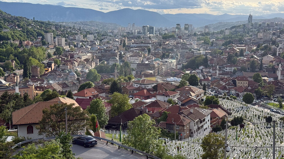 Blick über Sarajevo mit dicht bebauter Innenstadt, umliegenden Bergen und einem großen Friedhof im Vordergrund.